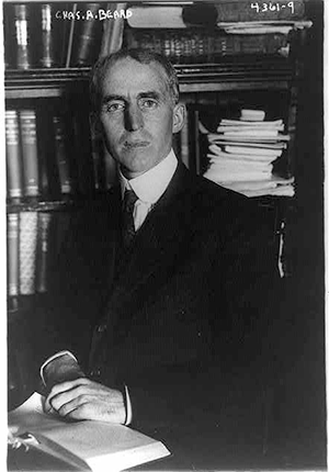 Charles A. Beard in front of bookshelf at table, half-length portrait, 1917.