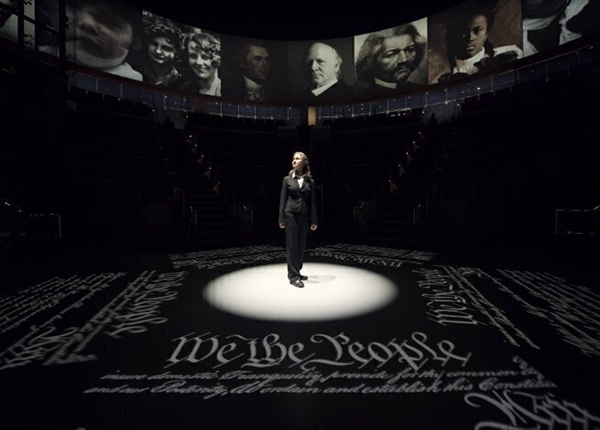 woman on stage with spotlight and text from the Declaration of Independence projected on the stage and photographs of historic figures above.