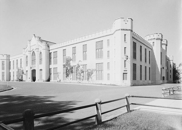 Two story building with ornate, castle-like outline and windows. Angled view showing front and side of the building with driveway and fenced grass in the foreground.